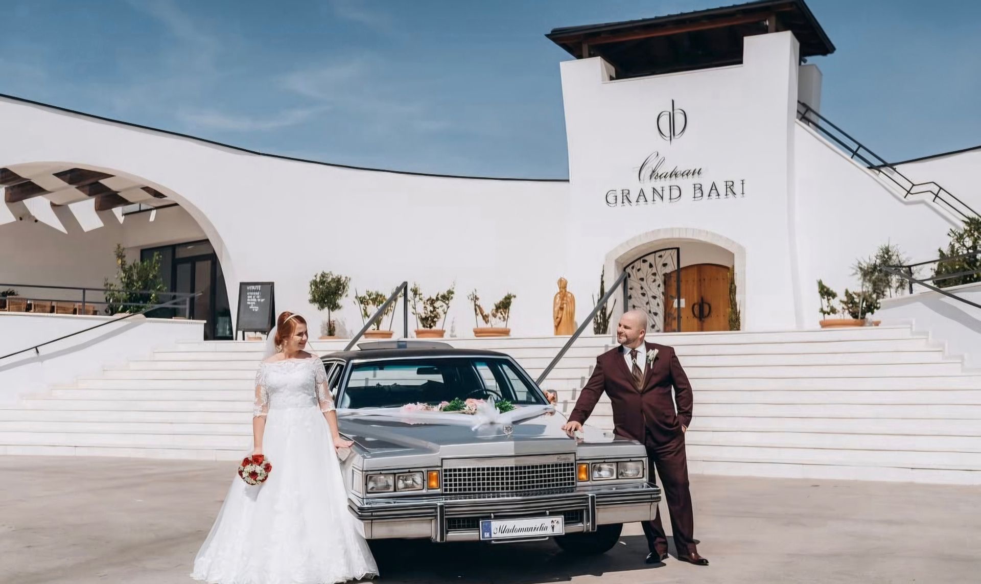 Nevesta a ženích po stranách prednej časti Cadillacu pred Chateau Grand Bari
